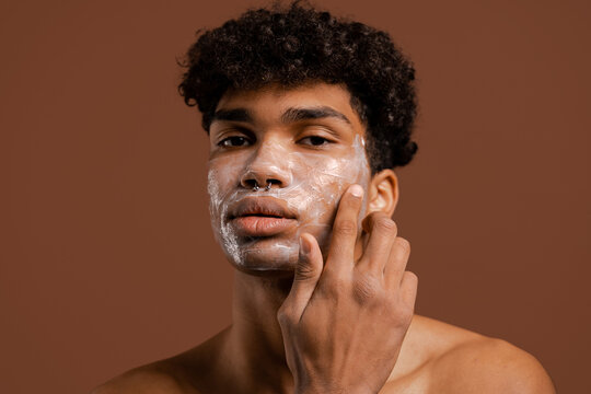Photo of attractive black man with piercing puts cream or nutrition mask on face. Naked torso, isolated brown color background