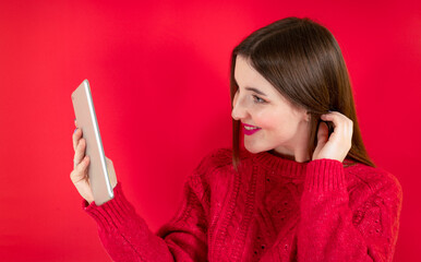 Naklejka premium Portrait of stunning joyful young woman in red fur sweater using tablet pc computer isolated on bright red wall background in studio