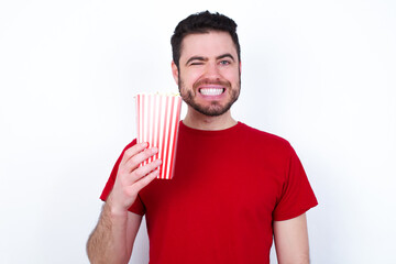 Coquettish Young handsome man in red T-shirt against white background eating popcorn smiling happily, blinking at camera in a playful manner, flirting with you.