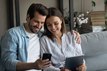 Millennial couple in love confident internet users sit on couch hold phone tablet exchange data share information between devices. Young husband wife send receive files app from cell to pad using wifi