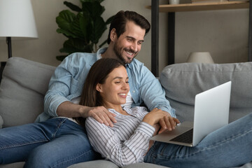 Smiling young wife girlfriend relax on couch in affectionate husband boyfriend embraces hold...