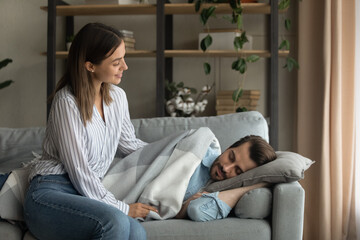 Obraz premium Love in details. Attentive millennial wife carefully covering tired husband sleeping on couch with warm soft plaid blanket. Affectionate young female take care of fatigued spouse resting on cozy sofa