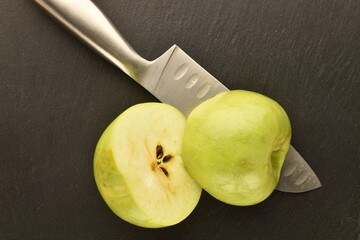 Two halves of a ripe green apple with a metal knife on a slate board, close-up, top view.