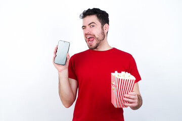 Young handsome man in red T-shirt against white background eating popcorn holding in hands cell...