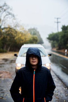 Young boy standing in front of car on side of road in rainstorm