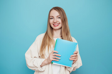 cheerful blonde student girl holding a book, notebook smiling, looking at the camera isolated on blue background in studio