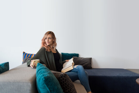 Woman sitting on couch with book and coffee looking away from camera