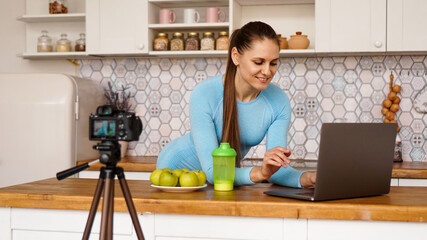 Fototapeta na wymiar Young woman in kitchen with laptop smiling. Food blogger concept. A woman is recording a video about healthy eating. Camera on a tripod.