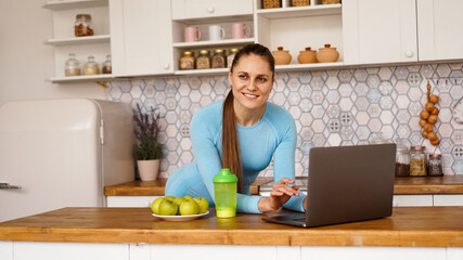 Fototapeta na wymiar Smiling woman using computer in modern kitchen interior. Cooking and healthy lifestyle concept. A woman is looking at the camera and smiling