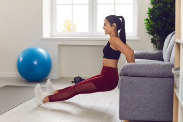 Fit female doing sports exercise for stronger arms at home, simply using her sofa. Side view of happy young woman doing triceps dips or reverse push-ups gripping edge of her couch in the living-room