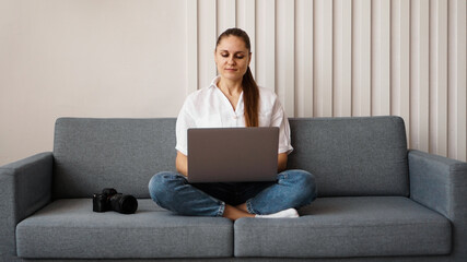 Fototapeta premium Happy young woman using laptop at home. On the couch next to the woman is a camera. Photographer retouches photographs at home.