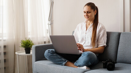 Happy young woman using laptop at home. On the couch next to the woman is a camera. Photographer...