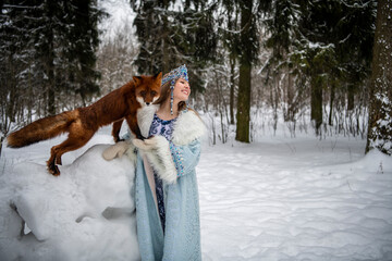 Russian beauty in a national costume with a red fox in a winter snowy forest 