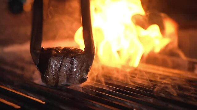 Cooked steak mignon is taken from a hot, burning grill. Metal tongs on a macro plan in slow motion