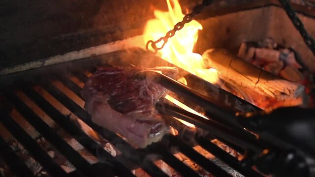 Grilled tibon steak flipped over with metal tongs in a black glove over a background of fire