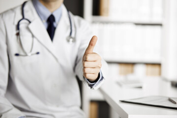 Unknown male doctor sitting with thumbs up sign at his working place in clinic, closeup. Perfect medical service in hospital. Medicine and healthcare concept
