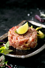 Tuna tartare with green salad, lime, avocado and mustard sauce serving on japanese style black ceramic plate over black marble background. Restaurant appetizer. Close up