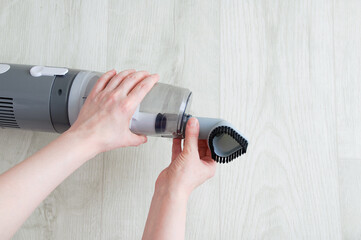 The hand of a caucasian woman puts on a small nozzle on a gray vacuum cleaner on white wooden background.