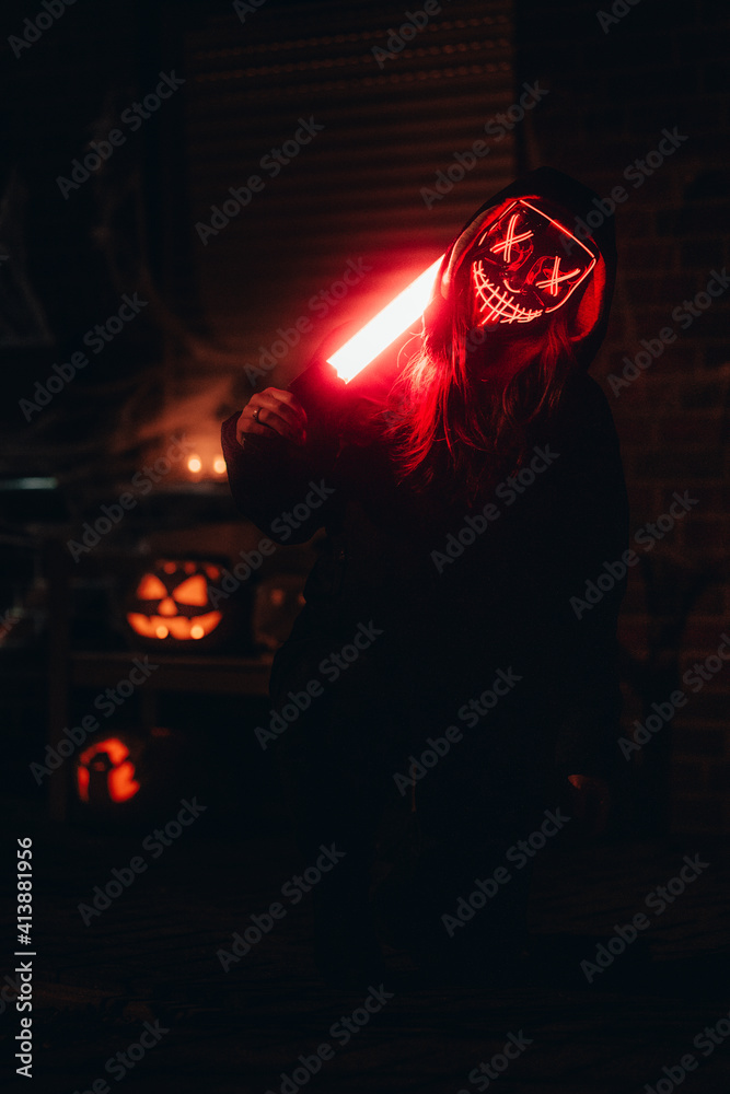 Wall mural closeup view of a girl with a red glowing dead mask with a lightsaber next to pumpkins on halloween