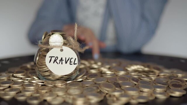 Blurred woman calculating stack of euro coins and putting them inside the glass jar with travel inscription. Female counting her monthly earnings, planning budget for summer vacations and travel