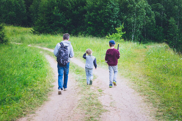 a man with his daughter and son are walking along the path in a summer forest during his hiking trip