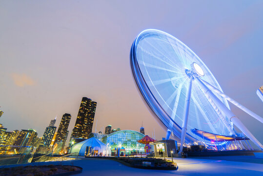 Illuminated spinning Ferris Wheel by modern buildings against sky in city during dusk