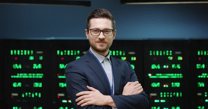 Successful male programmer in jacket and glasses standing on background of running servers looking at camera with his arms crossed.