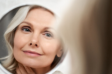 Happy 50s middle aged woman model touching face skin looking in mirror reflection. Smiling mature old lady pampering, healthy moisturized skin care, aging beauty, skincare treatment cosmetics concept. - obrazy, fototapety, plakaty