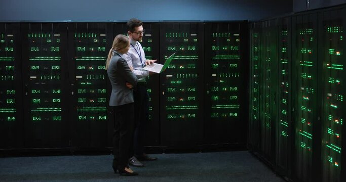 Young programmer holding laptop in his hands shows blonde woman manager the serviceability of computers in server room.