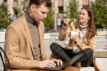 Young serious elegant man using laptop while his happy girlfriend eating wok