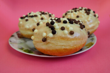 close up three round doughnuts in white frosting with chocolate balls in a white plate with flowers on a pink background side view