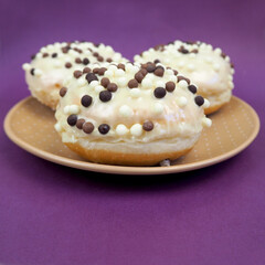 three round doughnuts in white frosting with chocolate balls in a brown plate on a lilac background side view