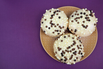 three round doughnuts in white frosting with chocolate balls in a brown plate on a lilac background top view