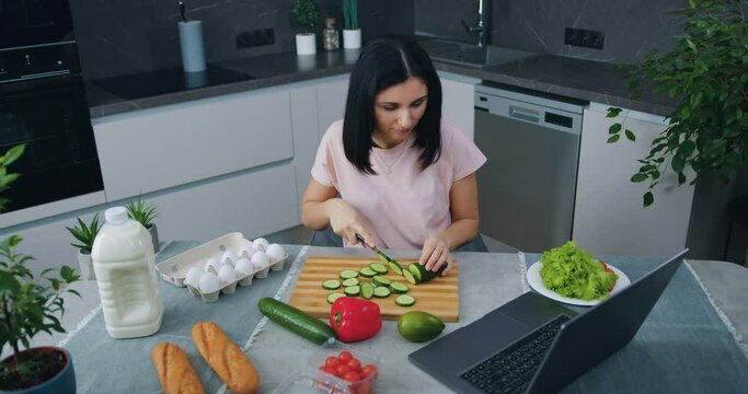 Pleasant calm smiling young brunette sitting at the table in modern cuisine and cutting cucumber for vegetable salad simultaneously choosing interesting program on laptop