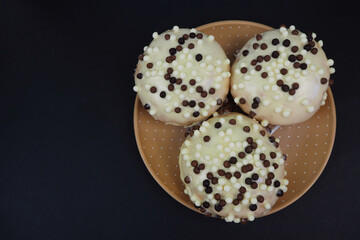 three round doughnuts in white frosting with chocolate balls in a brown plate on a black background top view