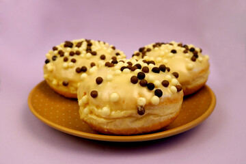 three round doughnuts in white frosting with chocolate balls in a brown plate on a purple background side view . high-calorie food