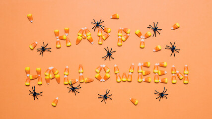 Overhead view of candy corns arranged as Happy Halloween text amidst artificial spiders over orange background