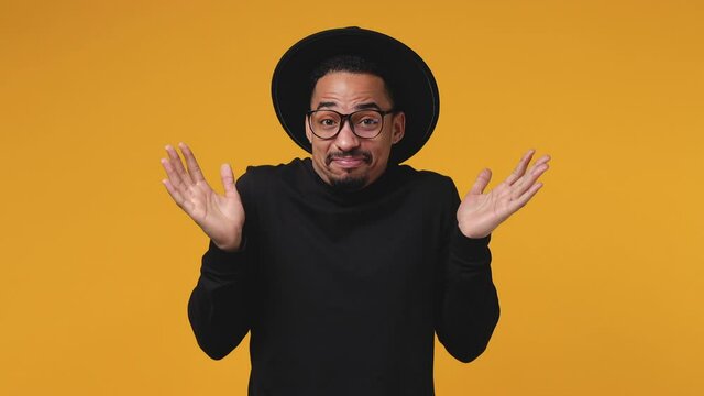 Confused puzzled young african american man 20s years old in casual black clothes hat eyeglasses isolated on yellow color background in studio. People lifestyle concept. Spreading hands say oops