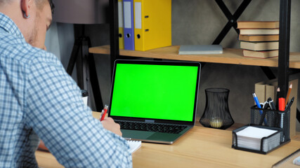 Green screen display laptop computer concept: Businessman at home office sitting on chair at desk writes information in notebook. Online remote distance business meeting webcam video conference call