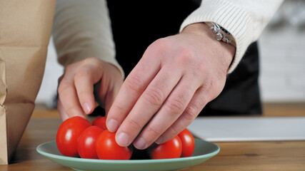 Professional chef man in black apron standing near table prepares ingredients vegetables for salad at home kitchen. Close up hands put fresh food red cherry tomato in plate