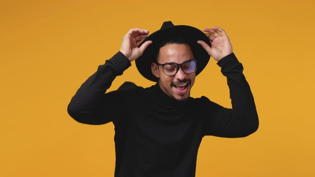 Funny african american man 20s in basic black clothes hat eyeglasses isolated on yellow background studio. People lifestyle concept. Dancing clenching fists waving rising hands point fingers sing song
