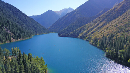 Kolsay lake among green hills and mountains. The mountain lake is surrounded by green forest, tall coniferous trees, grass and bushes. Clean water is like a mirror. Tourists swim on boats. Kazakhstan