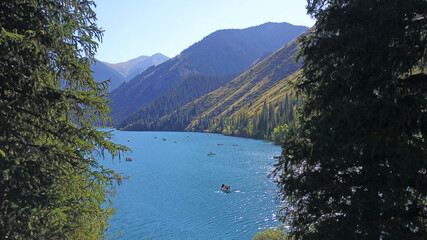 Kolsay lake among green hills and mountains. The mountain lake is surrounded by green forest, tall coniferous trees, grass and bushes. Clean water is like a mirror. Tourists swim on boats. Kazakhstan