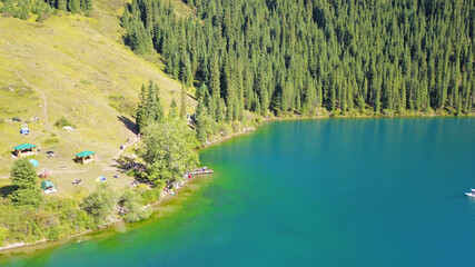 Kolsay lake among green hills and mountains. The mountain lake is surrounded by green forest, tall coniferous trees, grass and bushes. Clean water is like a mirror. Tourists swim on boats. Kazakhstan