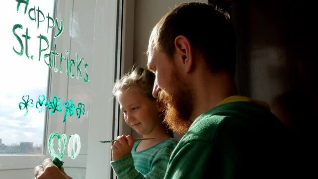 Drawing St. Patrick's Day Father with daughter painting green three-leaved shamrocks indoor, festive home decoration,family leisure. Drawing clover leaves on window glass. Stay home concept New normal