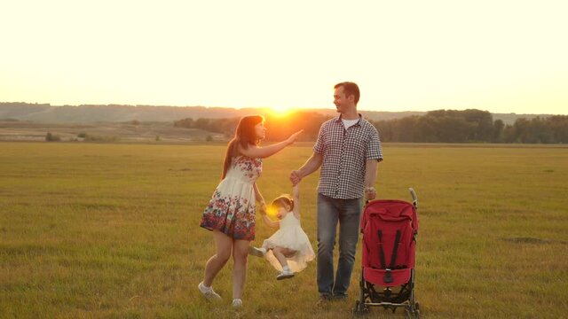 Happy family, child plays with dad and mom on field in light of sun. Little daughter plays and jumps holding hands of dad and mom in park at sunset. Walking with small child in nature, baby carriage