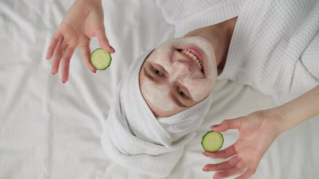 Skin Care, After Bath, Cosmetics at Home, Caucasian Woman, Anti Aging. Cheerful caucasian woman in a bathrobe with a towel on her head lies on the bed and uses a cosmetic face mask and cucumber