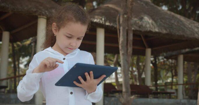 The child has a rest plays in the tablet outdoors. Curious cute girl 7-8 years old is having fun with a digital tablet device watching a cartoon while walking on the beach.