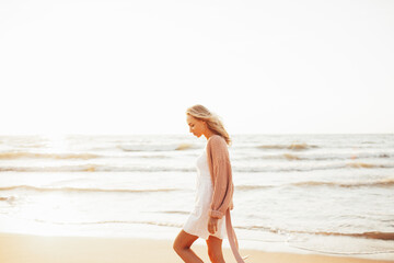 Young slim woman walk alone on the beach or ocean at summer sunset. Female dressed in a warm sweater.