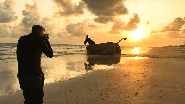 Man taking a photograph of a WWII bunker turned into a sculpture of a mule at sunset on the beach in Blavand, Denmark